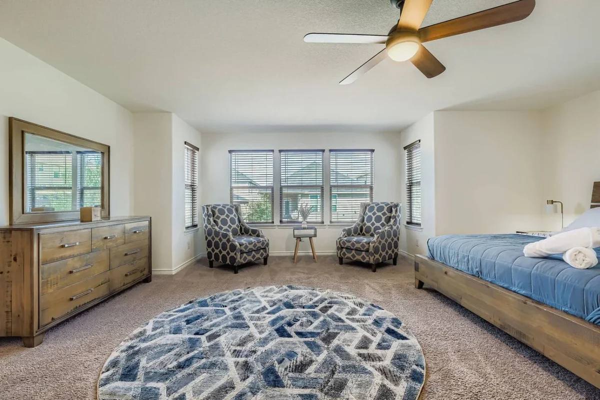 Bedroom with blue bedding, a wooden dresser, patterned chairs by the window and a ceiling fan